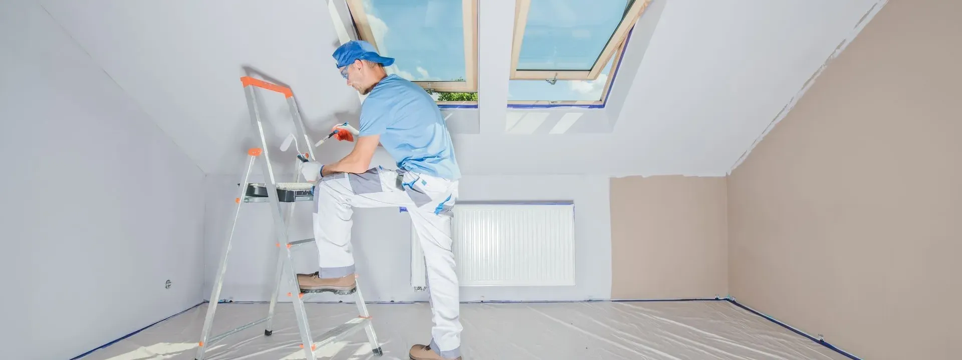 A painter on a stepladder in a bright attic room, the floor covered in plastic sheeting — interior painting in New Bedford, MA