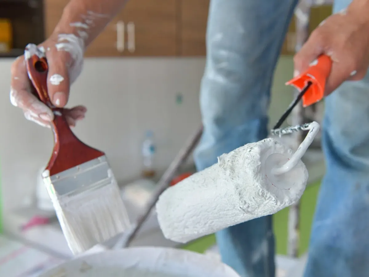 A painter's hands holding a brush and a roller, covered in white paint — prep for interior painting in New Bedford, MA
